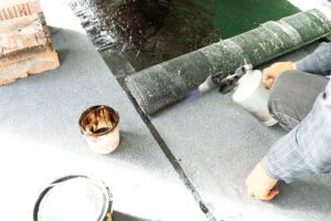 A person wearing orange gloves, likely a Flux - best-rated waterproofing specialist, applies a waterproofing membrane to a surface using a blowtorch; a can of adhesive is nearby.